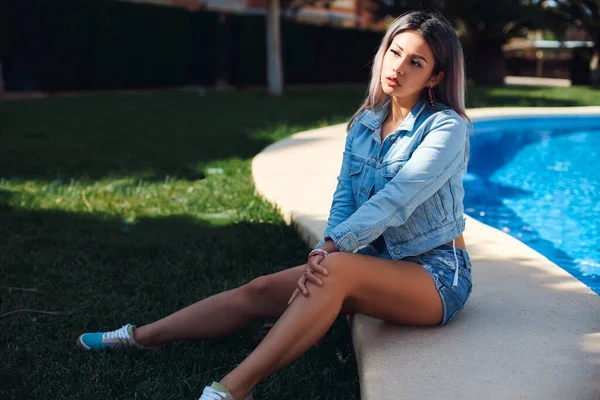 Güneşin altında havuz kenarında oturan güzel bir sarışın ve kot ceketli palmiye ağaçları ve şort ve açık bir mayo. İyi tatiller..