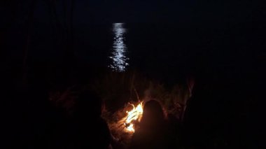 Bir grup insan ay yansıyan gece sahil üzerinde kamp olduğu deniz karşı ateş yakın