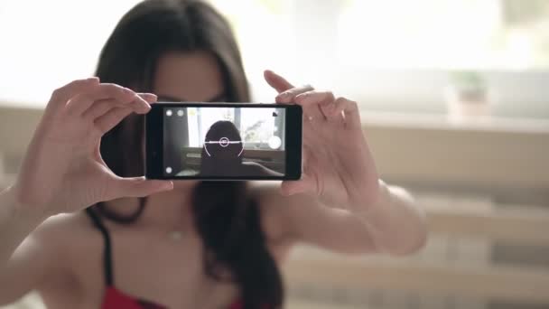 Schöne lächelnde brünette junge Frau in sexy roten Dessous macht Selfie mit ihrem Smartphone auf dem Bett sitzend — Stockvideo