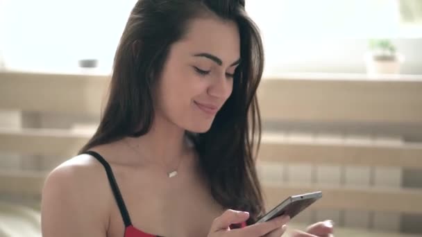 Красивая улыбающаяся брюнетка молодая женщина в сексуальном красном белье печатает на своем смартфоне сидя на кровати — стоковое видео