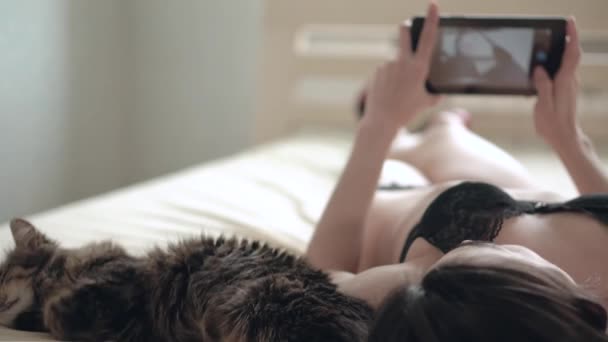 Joven hermosa mujer en lencería negra haciendo foto selfie en una tableta acostada en una cama cerca del gato — Vídeos de Stock