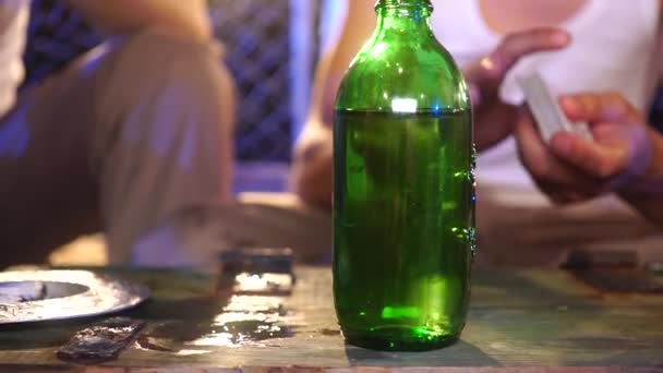 Mann im weißen T-Shirt in der Unschärfe hinter einer grünen Flasche mischt die Karten in seiner Hand, bevor er auf der alten Holzkiste spielt — Stockvideo