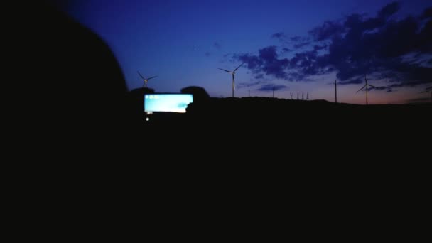Дівчата силует зйомки декількох на смартфоні Вітряки під час нічної сутінки на горизонтальній вітроенергетиці в полі з дивовижними хмарами вище — стокове відео
