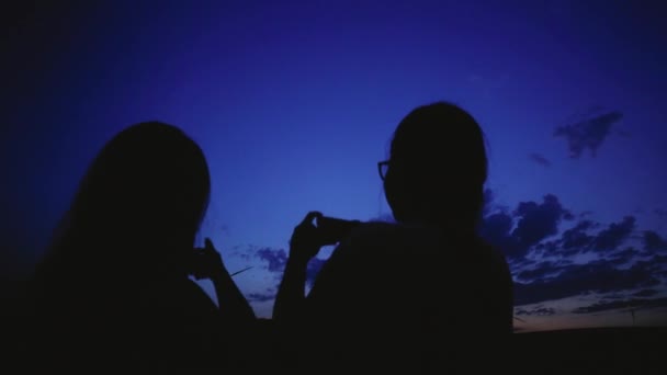 Duas meninas discutem moinhos de vento e tiram uma foto da paisagem noturna usando o smartphone — Vídeo de Stock