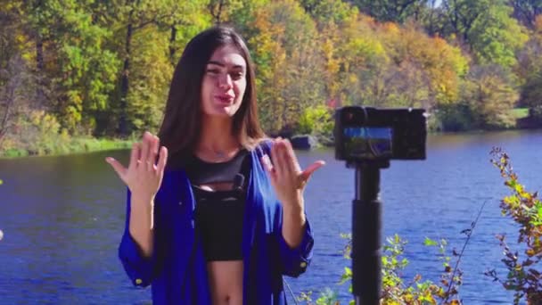 Брюнетка блоггер на берегу реки записывает видео для видеоблога с помощью камеры — стоковое видео