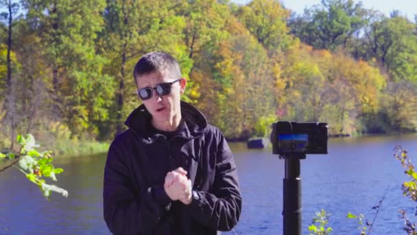 Morena hombre blogger en gafas de sol en la orilla del río está grabando vídeo para vlog utilizando la cámara — Vídeos de Stock