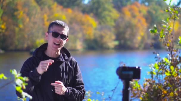 Bloguer jeune homme dans des lunettes de soleil debout à côté de la caméra à la nature montrant pouces levés et dire au revoir pour les abonnés au ralenti — Video