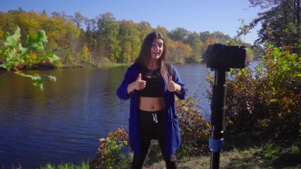 Блог молодая женщина стоять рядом с камерой на природе говорить и жесты замедленной съемки показывая большие пальцы вверх и улыбаясь — стоковое видео