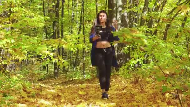 Blogger charismatic young woman is walking in autumn forest and recording video for vlog using camera talking actively gesturing and smiling — Stock Video