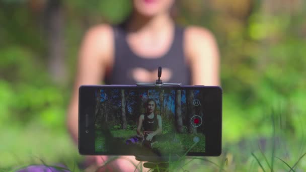 美しい若い女性のブロガーの話をサブスクライバーにスマート フォンを持つ公園で彼女のチャネルのビデオ トレーニングのプロセスを撮影 — ストック動画