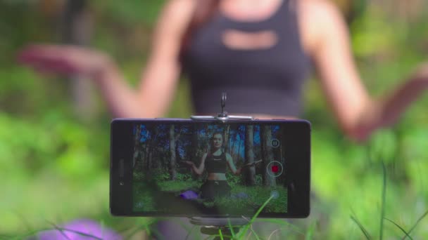 Красивая молодая женщина блоггер показывает дыхательные упражнения разговаривая с абонентами и съемки видео для своего канала в парке со смартфоном — стоковое видео
