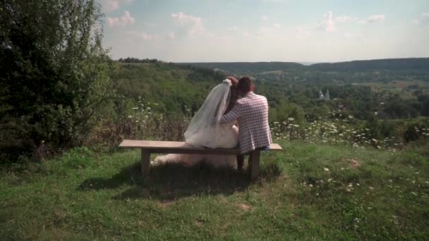 Romantické svatební pár sedí na lavičce a dívají se na církev — Stock video