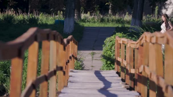 Junge Frau mit weißem Fächer im weißen Kleid geht an einem sonnigen heißen Tag auf die Holzbrücke — Stockvideo