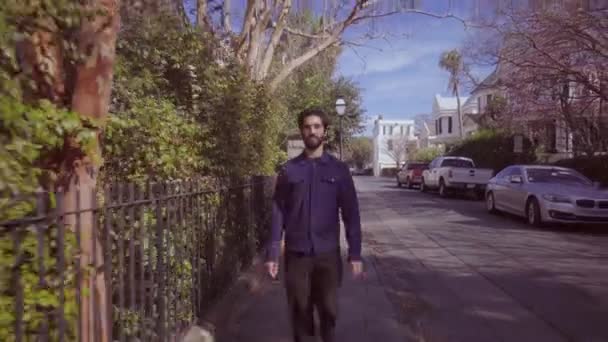 Молодой бородатый человек идет по улице вдоль изгороди — стоковое видео