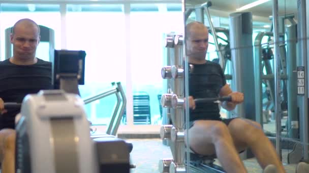 Young athlete makes an exercise on rowing simulator in gym reflecting in mirror — Stock Video