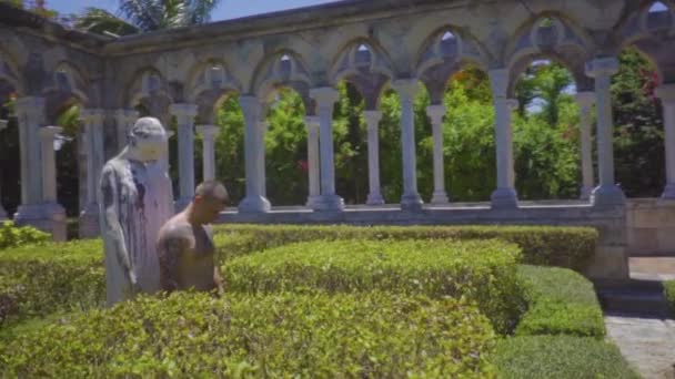 El hombre con tatuajes se para frente a una estatua entre columnas antiguas con la cabeza inclinada en oración — Vídeo de stock