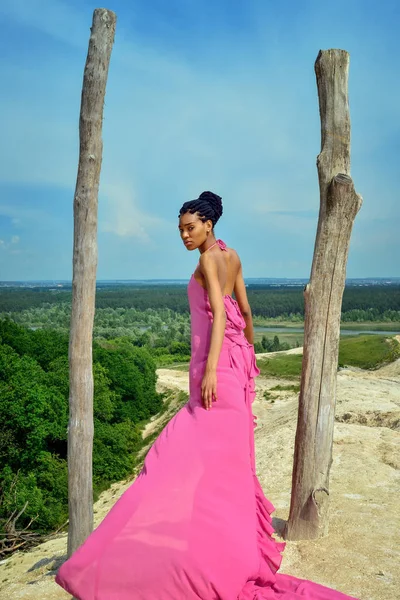 A beautiful African American girl, with dreadlocks, in a pink dress, stands on a hill between the dry trees and looks over her shoulder at the camera. Rear view from the back. Portrait. Vertical view