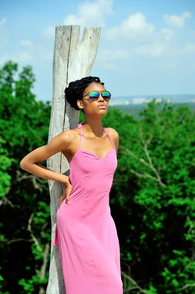 Pembe elbiseli, başında, güneş gözlüğü ve güneşe bakarak dreadlocks ile kırık bir kuru ağaç yakınındaki bir Afrikalı-Amerikalı kız duruyor. Portre
