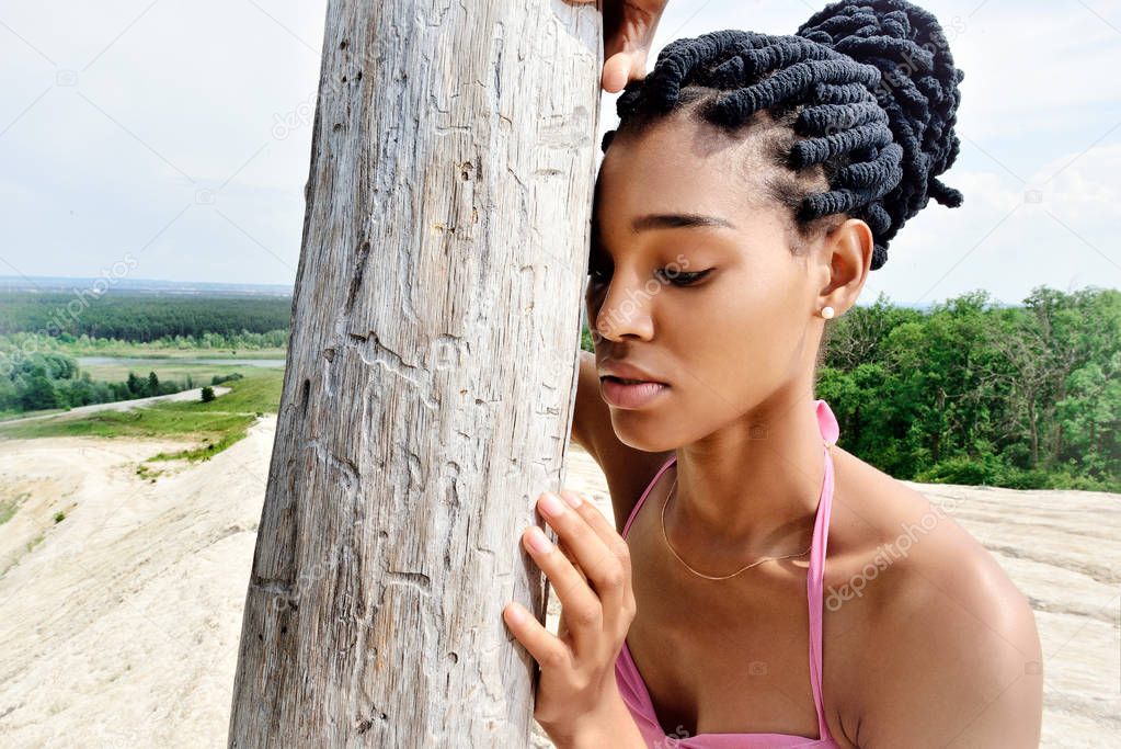 A beautiful African girl model, with dreads on her head, stands on a hill and thoughtfully embraces a dry tree. Portrait. Close up. Horizontal view