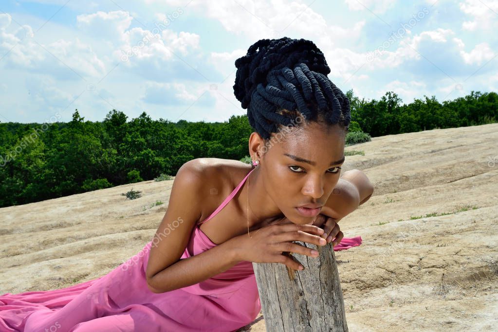 Sad African American girl lying in a pink dress on the mountain against the sky and clouds, leaning his hands on a dry tree and squinting looks at the camera. Portrait. Close up. Horizontal view