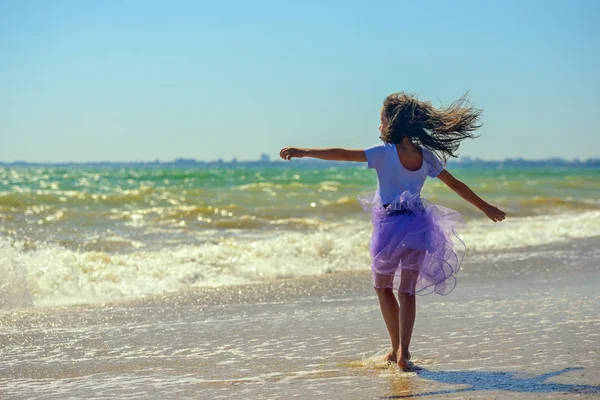 소녀는 파도의 배경에 보라색 드레스에 화창한 해변에서 그녀의 머리는 바람에 — 스톡 사진