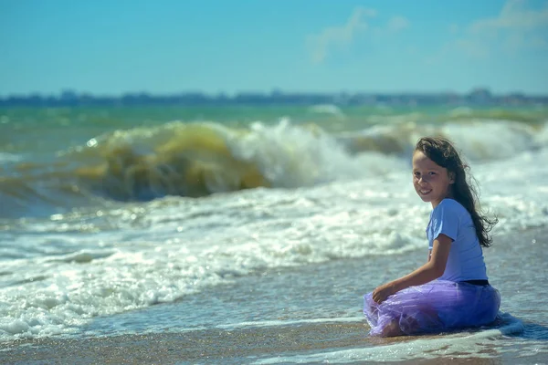 보라색 드레스에 해변에 가장자리에 그녀의 무릎에 카메라와 파도의 배경에 미소에 — 스톡 사진