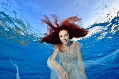 Kızıl saçlı beyaz bir elbise içinde geliştirme ile gelini su altında mavi bir arka plan üzerinde yüzen, kamera ve gülümsüyor görünüyor. Portre. Su altından alttan görünüm.
