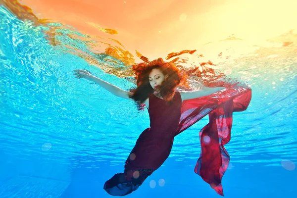 赤い髪と赤いドレスで美しい少女は 水泳や黄色の光と熱帯の夕日の背景にプールで水中ダンスします 肖像画 — ストック写真