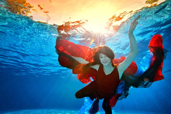 A playful girl with red hair swims underwater in the pool with red and blue fabric. She looks at the camera and smiles. Counter light from the sun. Portrait. Landscape image orientation.