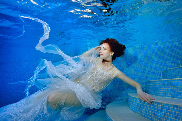 A beautiful girl in a transparent white dress with red hair swims underwater near the bottom of the pool and looks up. Portrait. Landscape orientation.