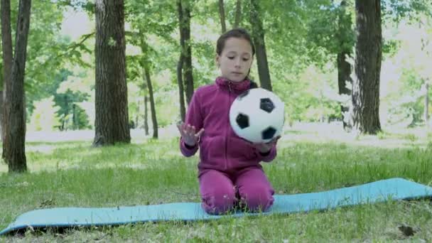 夏季には緑の芝生の公園で座っている 黒と白のサッカー ボールを保持して 彼と一緒に遊んでピンク トラック スーツの少女 Fps — ストック動画