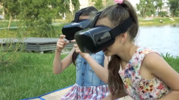 Dvě holčičky ve virtuální realitě přilby hraje v parku na trávě u řeky při západu slunce. Zblízka. Boční pohled. 4k, 29,97 fps.
