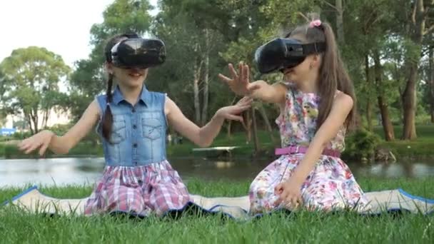 快乐的小女孩坐在公园的草地上 在河边的虚拟现实眼镜和玩虚拟游戏 他们挥动双手大笑 Fps — 图库视频影像