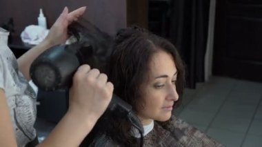 Güzel bir doğulu kız güzellik salonunda oturuyor ve ıslak saçlarını bir saç kurutma makinesi bulunan bir saç stilisti kurur. Saç Bakımı. Yan görünümü. 4 k. 25 fps.