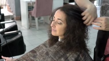 Güzel bir doğulu kız güzellik salonu, kamera ve gülümsüyor, bakar oturuyor ve Kuaför stilist onun saç şekillendirme yapar. Yakın çekim. 4 k. 25 fps.