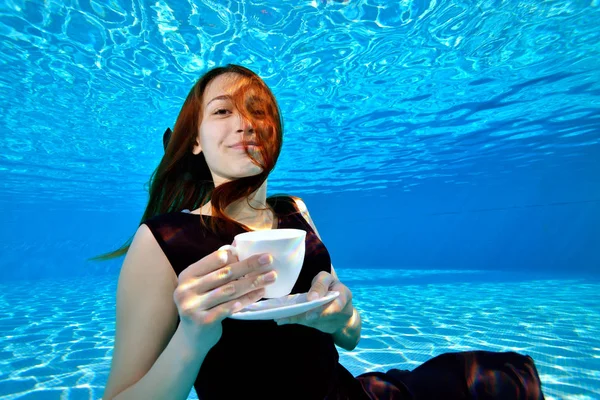 Beautiful young girl with red hair posing underwater in the pool with a white coffee Cup in her hands, looking at the camera and smiling. Portrait. Underwater photography. Landscape orientation.