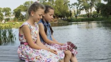 İki neşeli küçük kız bir ahşap köprü üzerinde yan yana Park City nehrin yaz aylarında oturabilirsiniz, yan ve gülmek için sabırsızlanıyoruz. Portre. Yakın çekim. 4 k. 29,97 fps.