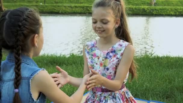 幸せな二人の少女は互いの反対の川での芝生の上に座って 自分の手で再生します 彼らは笑いながら手をたたいてごらん Fps — ストック動画