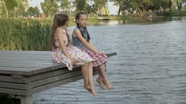 İki mutlu küçük kız çıplak ayakları ile yan yana Park City nehir kıyısında ahşap köprüdeki yaz aylarında su üzerinde oturuyor. Onlar kamera ve gülmek bak.