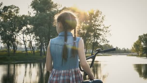 夕暮れ市公園の水の近くの自転車で立っているおさげのかわいい女の子 彼女は回る カメラを見て 優しく微笑みかけます クローズ アップ リアビュー Fps — ストック動画