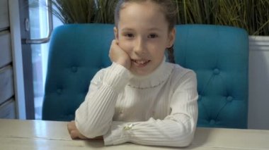 Kafasını ellerinin üzerinde koyarak ve gülümseyerek kameraya bakarak büyüleyici küçük bir kız ile beyaz bir elbise içinde ahşap bir masa, bir kafede otururken pigtails, yüzünde çiller ile. Portre.