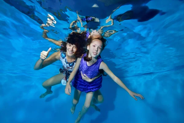 Underwater photography. Happy mom and daughter are swimming and playing underwater in the pool in beautiful dresses. They hug, look at the camera and smile. Portrait. Horizontal orientation.