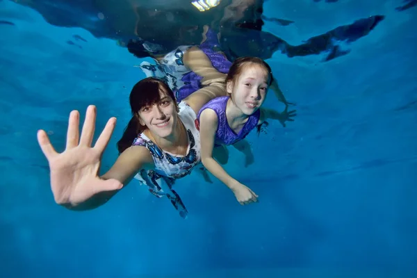 Happy mom with a little daughter in a purple dress floating under water in the pool. She extended her hand forward. They hug, look at the camera and smile. Portrait. Underwater photography.