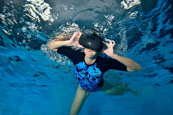 A cheerful girl plays underwater in the pool with black virtual reality glasses on her head. Virtual reality simulator. Shooting underwater. Horizontal orientation.