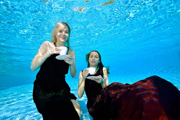 金发女郎和褐发女郎游泳 在水下的水池中与白色咖啡杯在他们手中的蓝色背景 他们看着相机笑了起来 水下摄影 — 图库照片