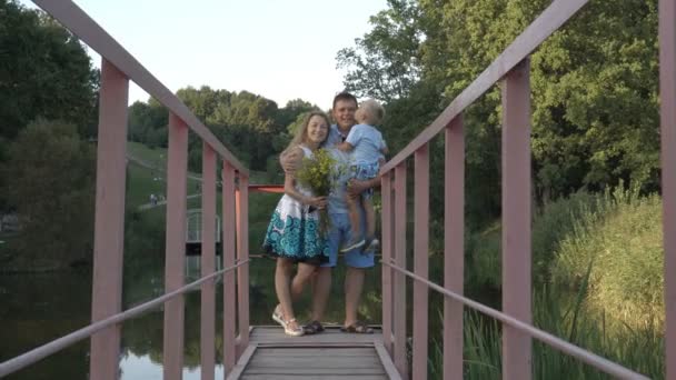 幸せな風景に対して公園の池の上の橋の上のお父さん お母さんと幼い息子が立っています 息子は教皇の手に座っている キスし 抱擁彼を彼の母と 生のビデオ — ストック動画