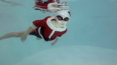 Küçük bir çocuk bir antrenör havuzu ile spor için Noel Baba gider gibi giyinmiş. O su altında merdivenlerden gözlük yüzme içinde yüzüyor ve kameraya doğru görünüyor. Ağır çekim. Sualtı çekim.
