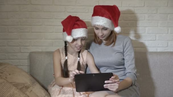 Дети в шляпах Санта-Клаус сидят вместе на диване, держа планшет, смотрят на него и улыбаются. Покупки в виртуальном магазине. 4К. 25 кадров в секунду . — стоковое видео