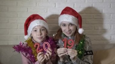 İki mutlu küçük kız kamera değerler 2019 gösteriyor. Otur ve bir beyaz tuğla duvar ve gülümseme karşı kanepede Santas şapkalı dans. Yakın çekim. 4 k. 25 fps.