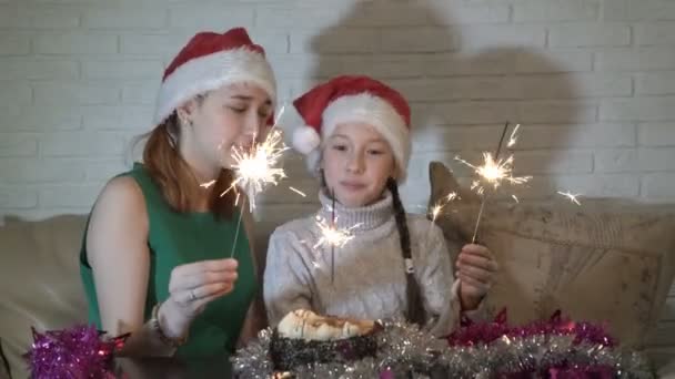 幸福的家庭 两个女孩 小和一个十几岁的圣诞老人的帽子与点燃火花在蛋糕坐在沙发上 看着他 面带微笑 圣诞节 闭上你的嘴4K — 图库视频影像
